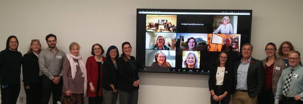 A photograph taken at the end of the SingWell 2023 Symposium of the in-person attendees gathered around a television displaying the online attendees participating over Zoom.